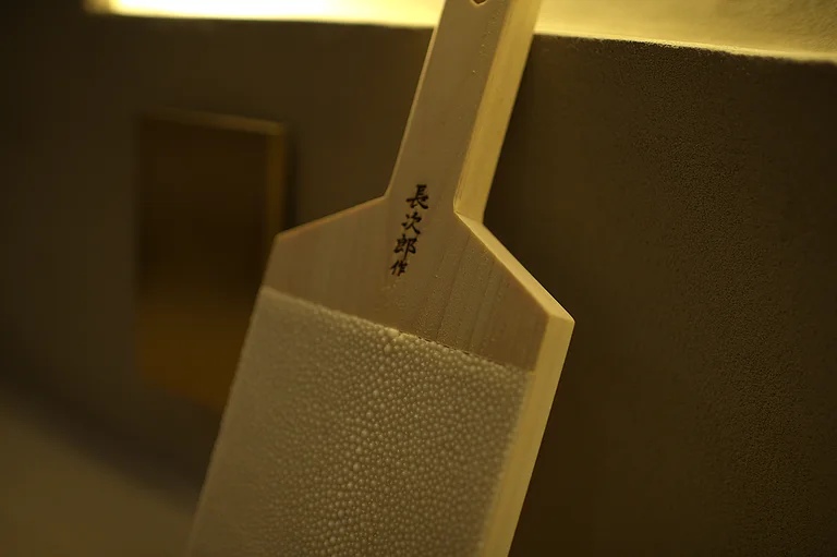 A wooden wasabi grater faced with sharkskin, hanging against the plaster wall